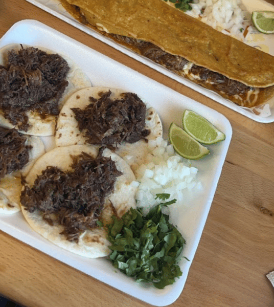 Mini tacos filled with shredded birria beef, served with diced onions, fresh cilantro, and lime wedges on a foam tray.