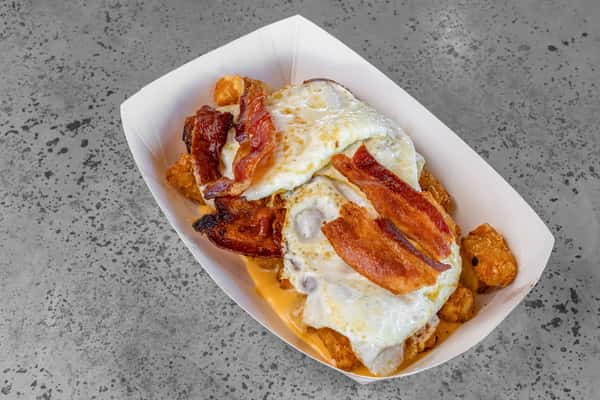Tater tots topped with melted cheese, crispy bacon strips, and over-easy eggs, served in a paper tray — a satisfying breakfast dish with classic diner flavors.