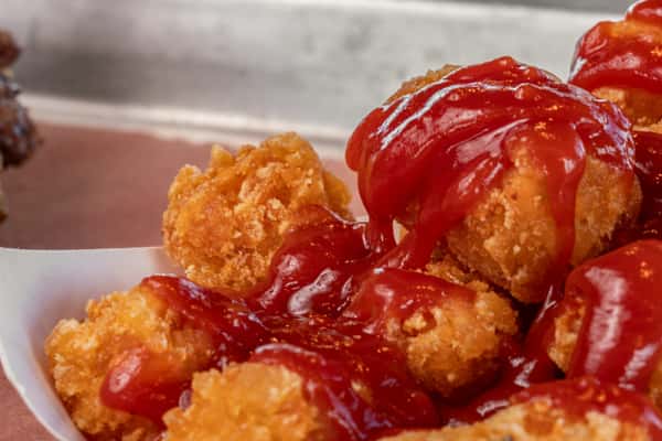 Close-up of crispy tater tots generously topped with ketchup, served in a paper tray — a popular side dish or snack ideal for fast-casual and diner-style menus.