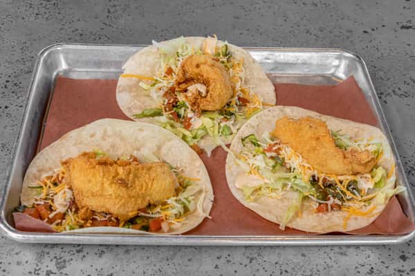 Three crispy fried fish tacos on soft flour tortillas, topped with shredded lettuce, diced tomatoes, shredded cheese, and creamy sauce — a Tex-Mex favorite served on a metal tray.