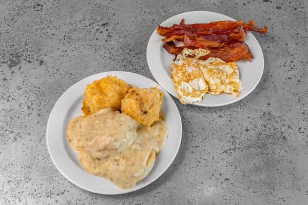 Southern-style breakfast with flaky biscuits smothered in creamy sausage gravy, crispy hash browns, over-easy eggs, and a generous side of crunchy bacon — hearty American diner favorite