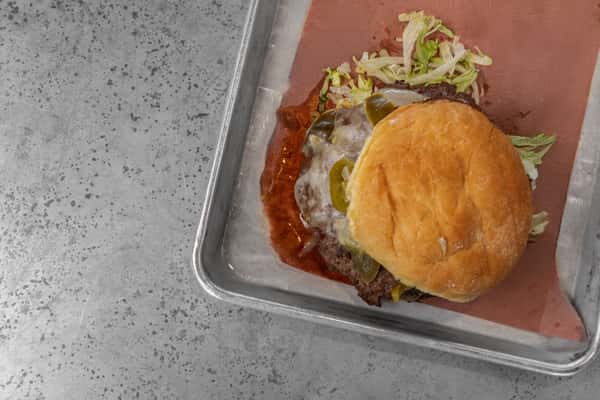 Cheeseburger topped with melted cheese, sliced jalapeños, and shredded lettuce on a toasted bun, served on parchment paper in a metal tray — a bold and spicy twist on a classic American burger.