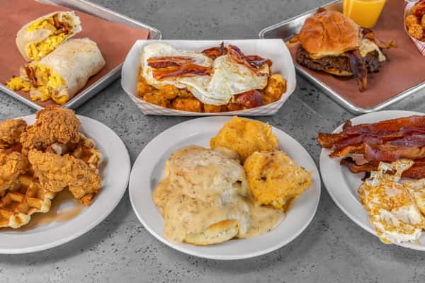 Variety breakfast platter featuring a burrito, bacon and eggs, biscuits with gravy, cheesy grits, chicken and waffles, and a bacon cheeseburger — ideal for showcasing a full Southern-inspired breakfast menu.