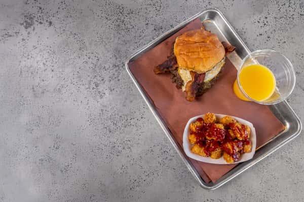 Hearty breakfast tray featuring a bacon cheeseburger with a fried egg, crispy tater tots drizzled with ketchup, and a fresh cup of orange juice — satisfying start to the day.