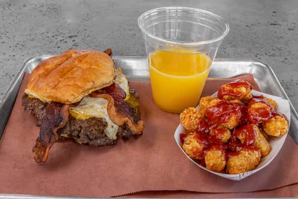 Breakfast-style bacon cheeseburger with a fried egg, served with crispy tater tots topped with ketchup and a cup of orange juice — hearty all-American breakfast platter.