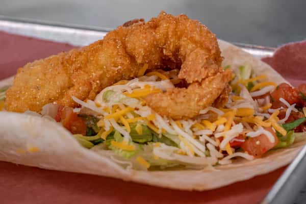 Close-up of a crispy fried shrimp taco topped with shredded lettuce, pico de gallo, and shredded cheese in a soft flour tortilla — a coastal twist on a Tex-Mex favorite.