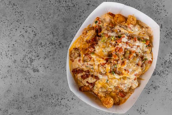 Loaded tater tots smothered in melted cheese, diced tomatoes, green onions, and creamy sauce, served in a paper tray — perfect for indulgent snacking or shareable appetizers.