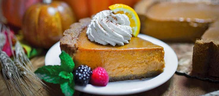 Slice of pumpkin pie with dollop of whipped cream with orange slice garnish and raspberry and blackberry garnish.