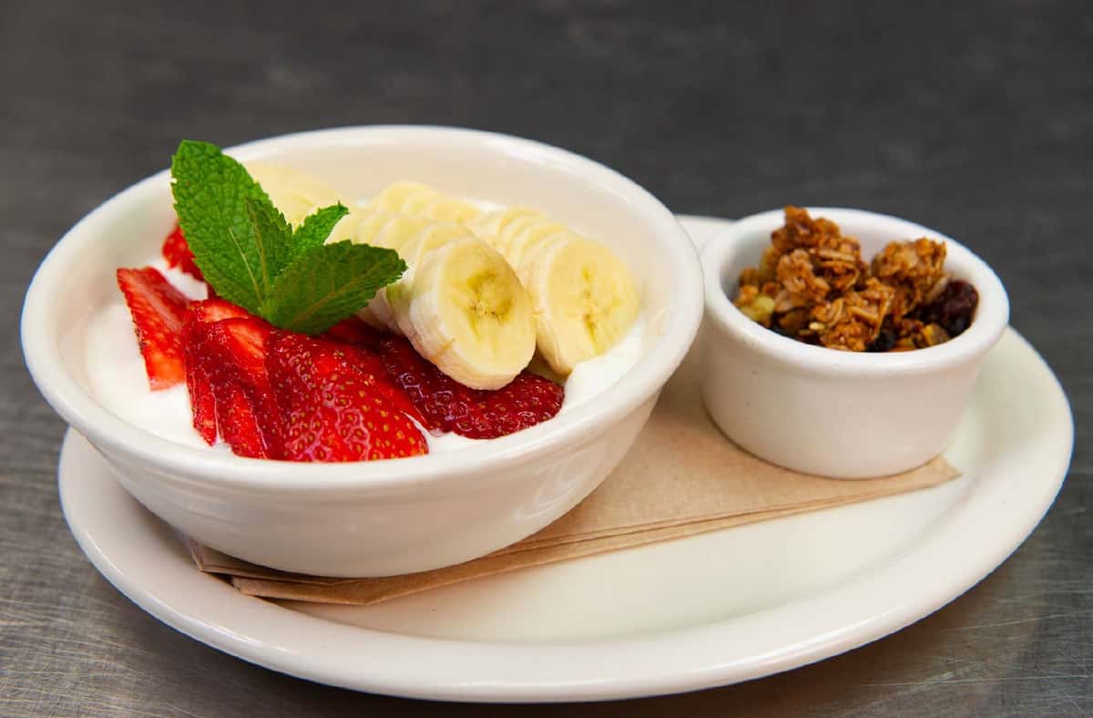 Fruit bowl with yogurt, sliced strawberries and bananas in which bowl with smaller white bowl with granola
