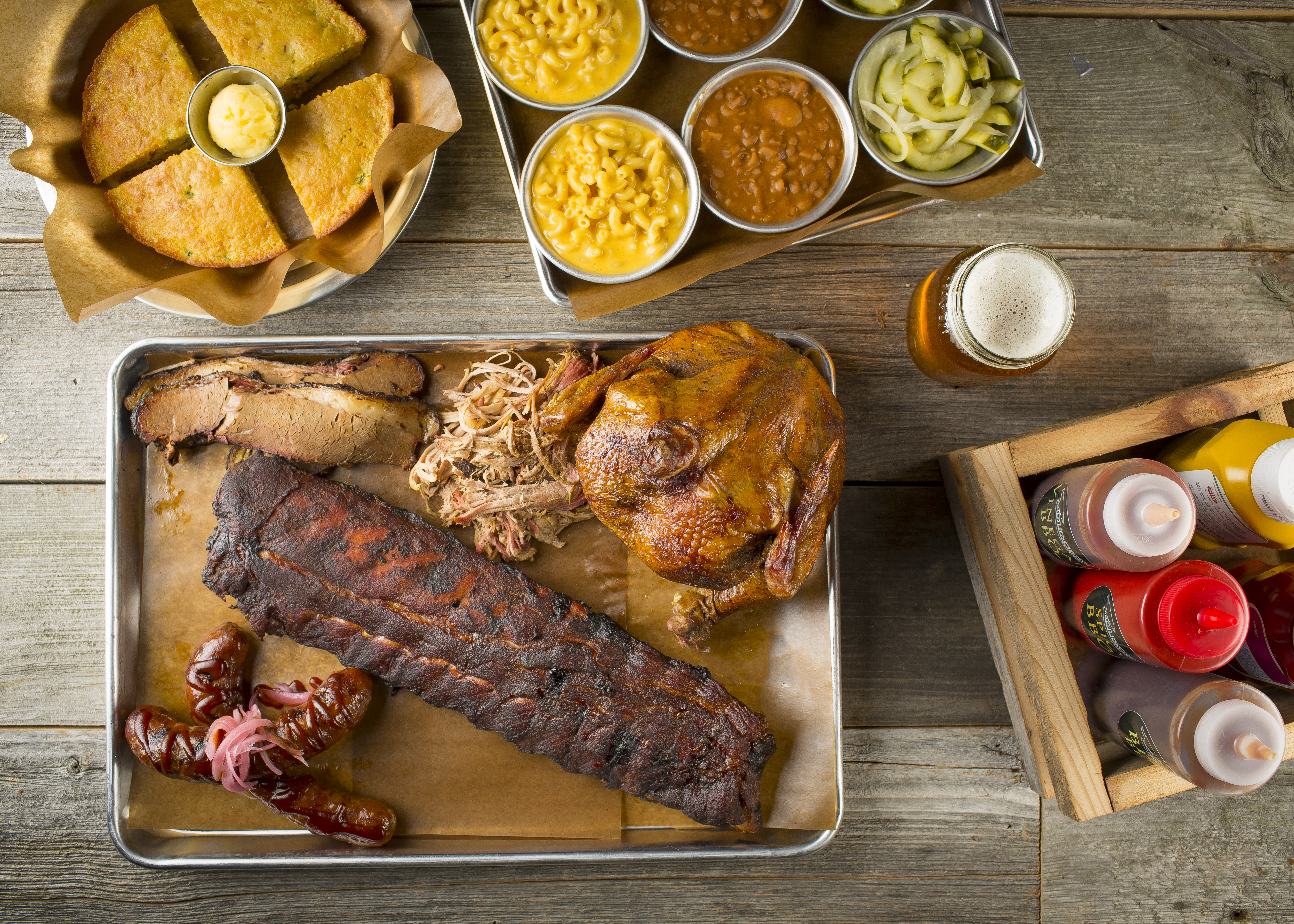 A platter of BBQ meats including ribs, pulled pork, chicken, and sausages, with sides of mac & cheese, baked beans, cornbread, and pickles.